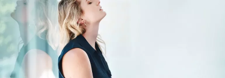 Woman in blue top leaning against glass wall, eyes closed.